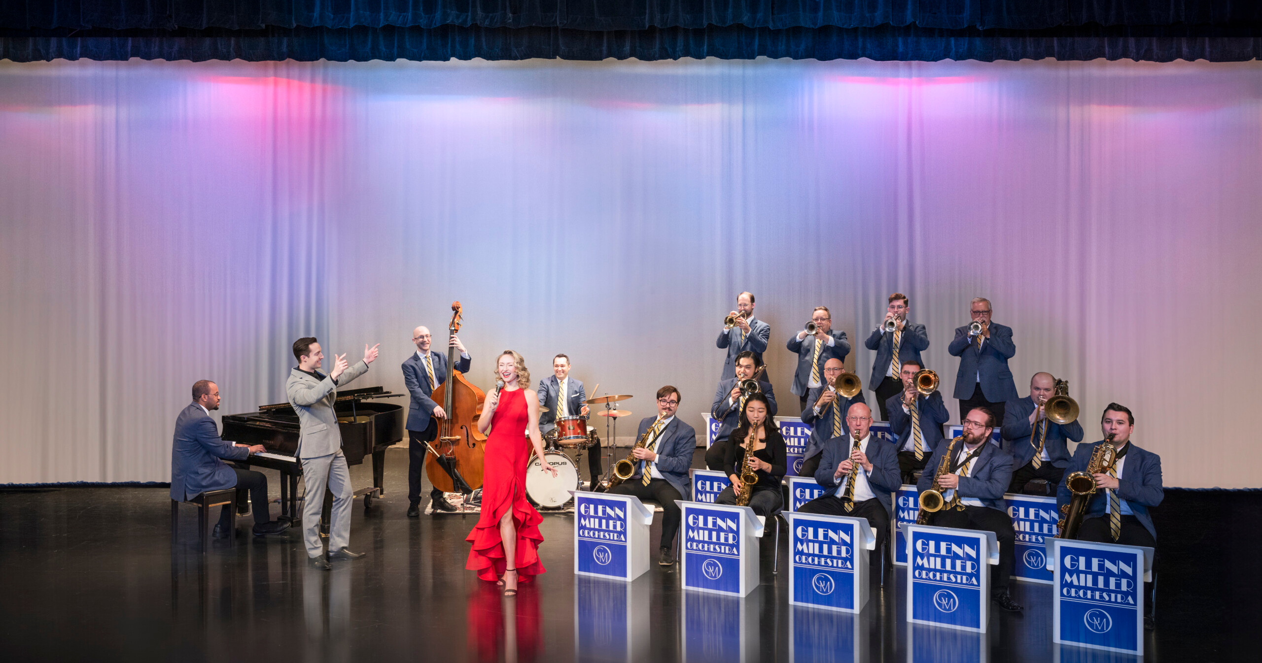 Big band jazz ensemble performs on stage, featuring musicians in blue suits and a vocalist in a red dress.